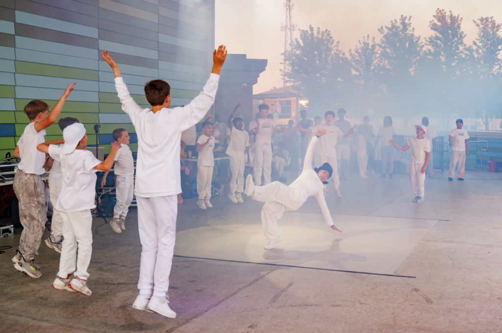 Group of children in white outfits performing a breakdance routine on an outdoor stage, with peers cheering in the background and a colorful wall to the left. A hazy blue smoke fills the right side of the scene.