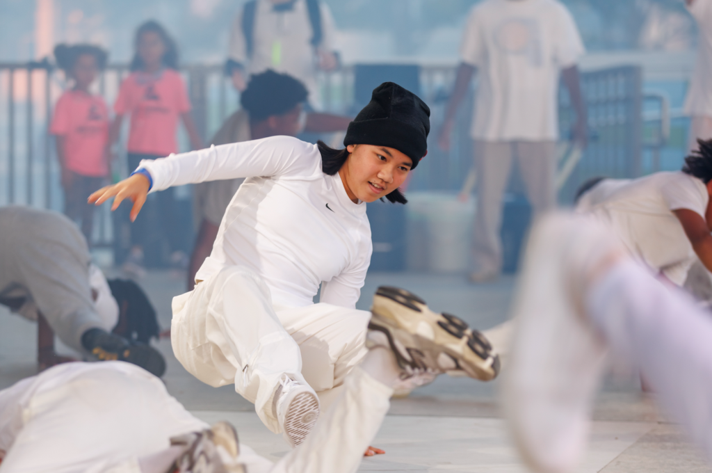 Dancer in white outfit and black beanie performing a mid-air breakdance move while onlookers watch in the background.