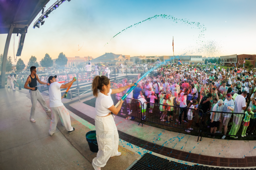 Performers on stage spraying colored powder toward a large crowd at a Holi-style festival outdoors.