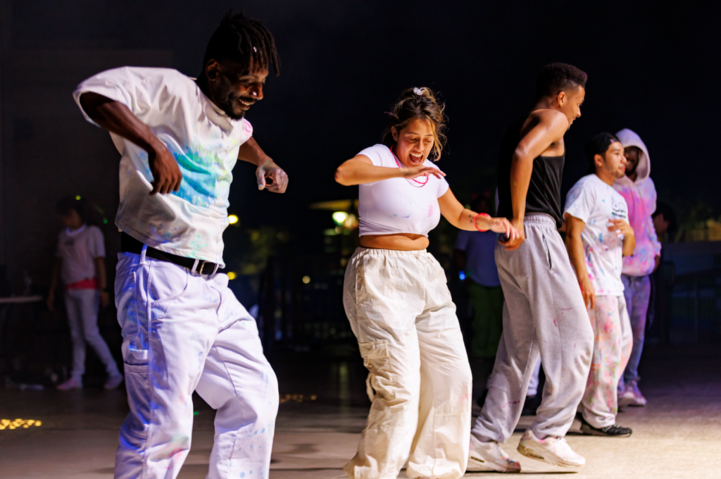 Group of young dancers in paint-splattered white clothes performing on a dimly lit stage at night, smiling and moving to the music.