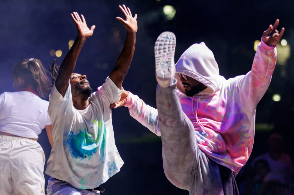 Two dancers raise their arms and kick high during a nighttime performance, wearing colorful, paint-splashed outfits.