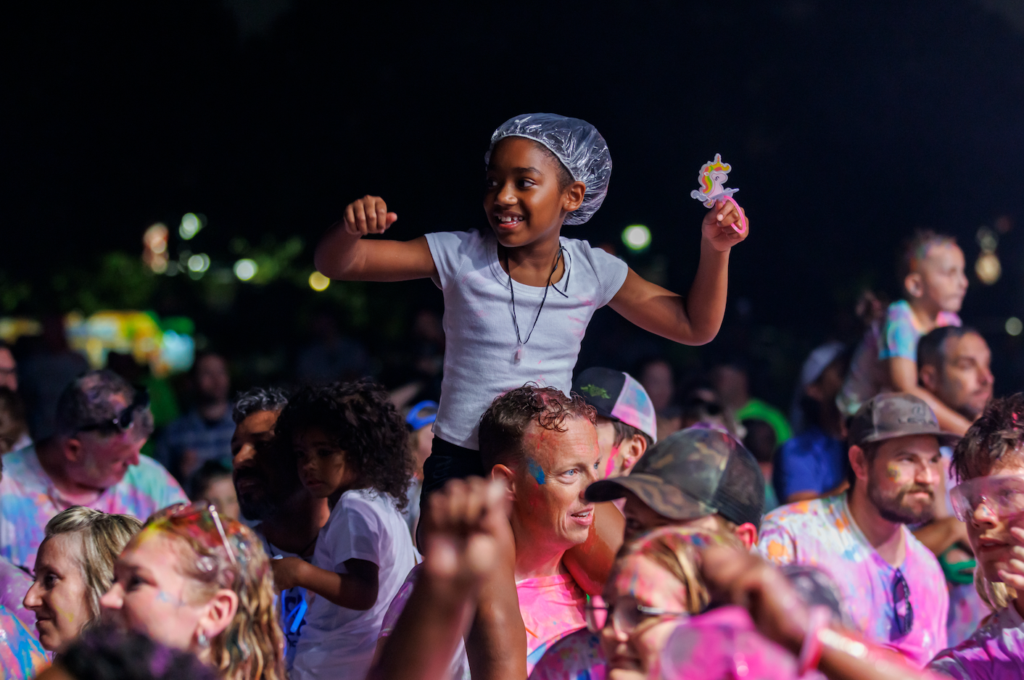 Child on someone's shoulders at a nighttime festival, cheering with a pink unicorn wand in hand amid a colorful crowd.