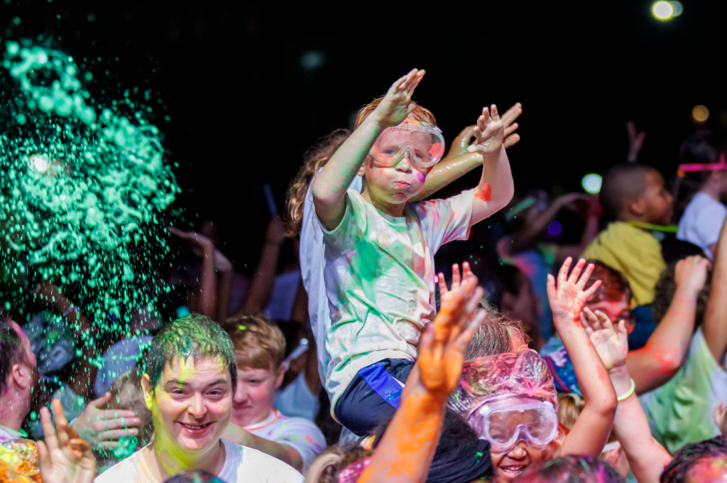 Child wearing goggles perched on another's shoulders, throwing colorful powder at a night festival.