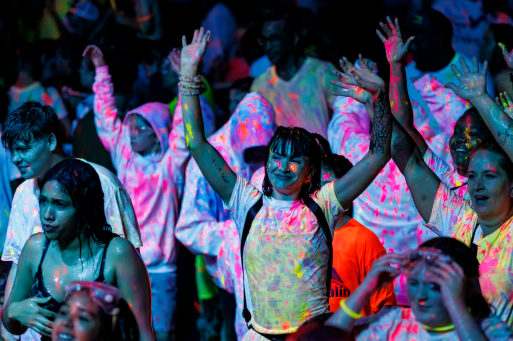 Crowd at a color run with glowing neon powder on clothes and skin, many hands raised in celebration at night
