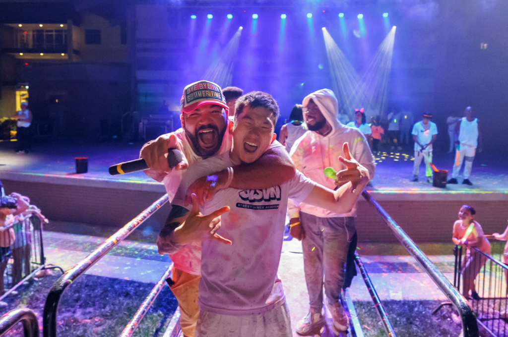Two men hug and pose with a microphone on a colorfully lit stage at a party in progress.