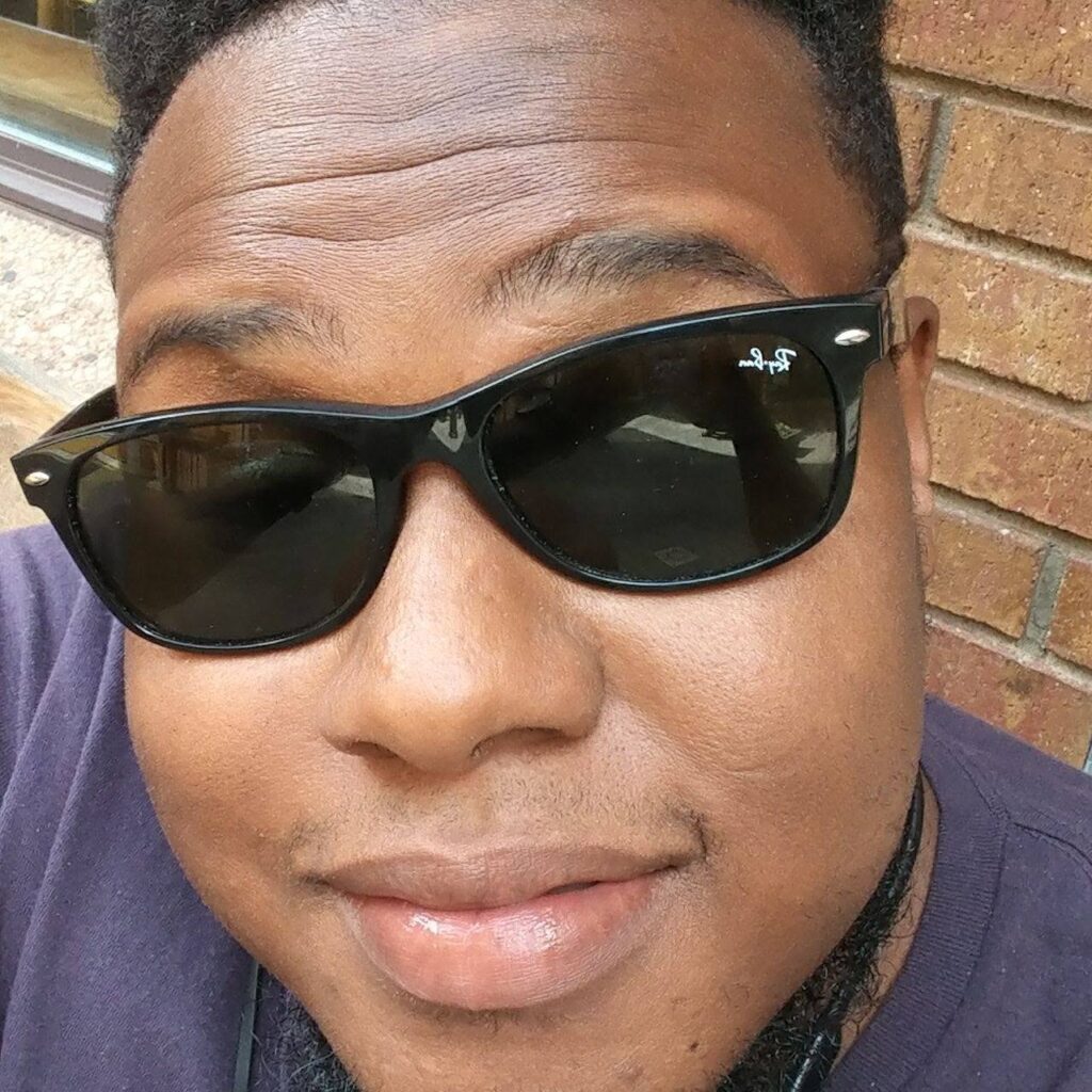 Close-up portrait of a person wearing black Ray-Ban sunglasses, smiling slightly with a brick wall in the background.